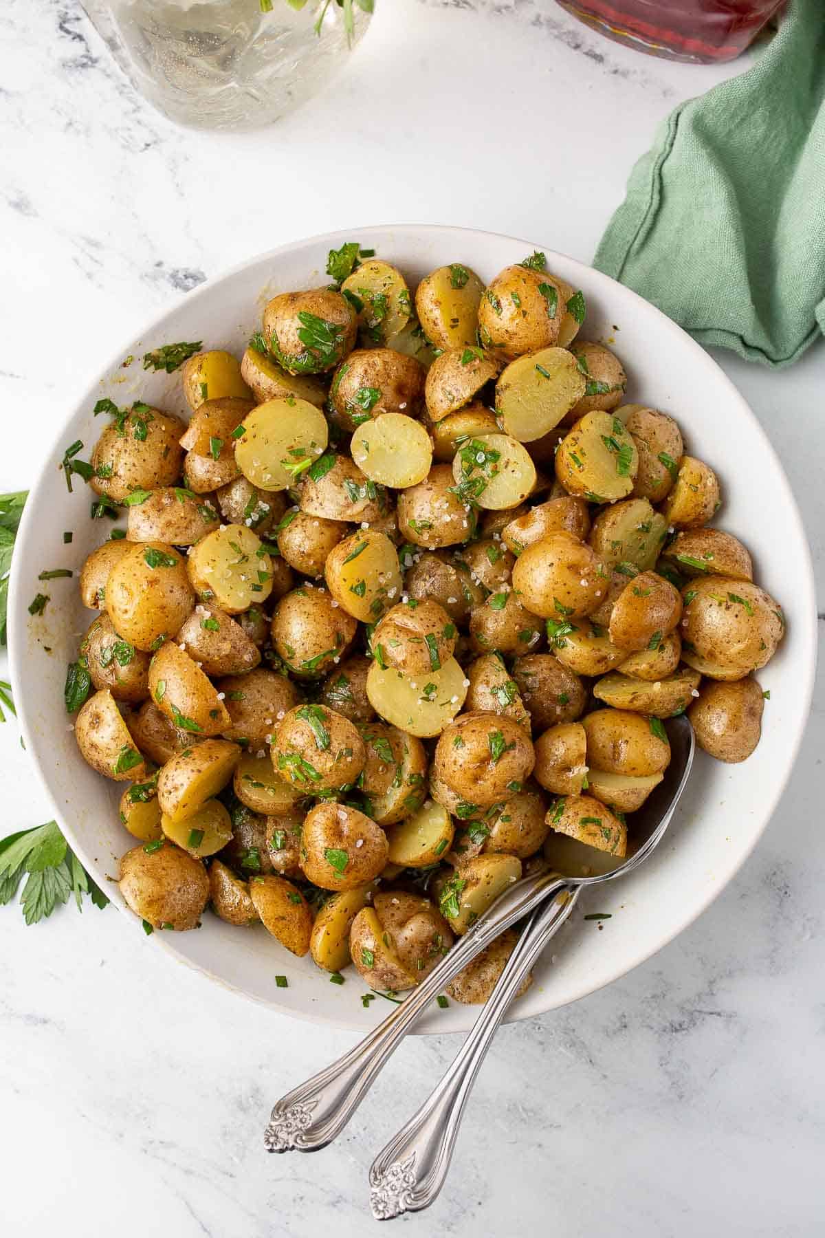 Overhead shot of German potato salad with herbs and utencils in large white bowl.