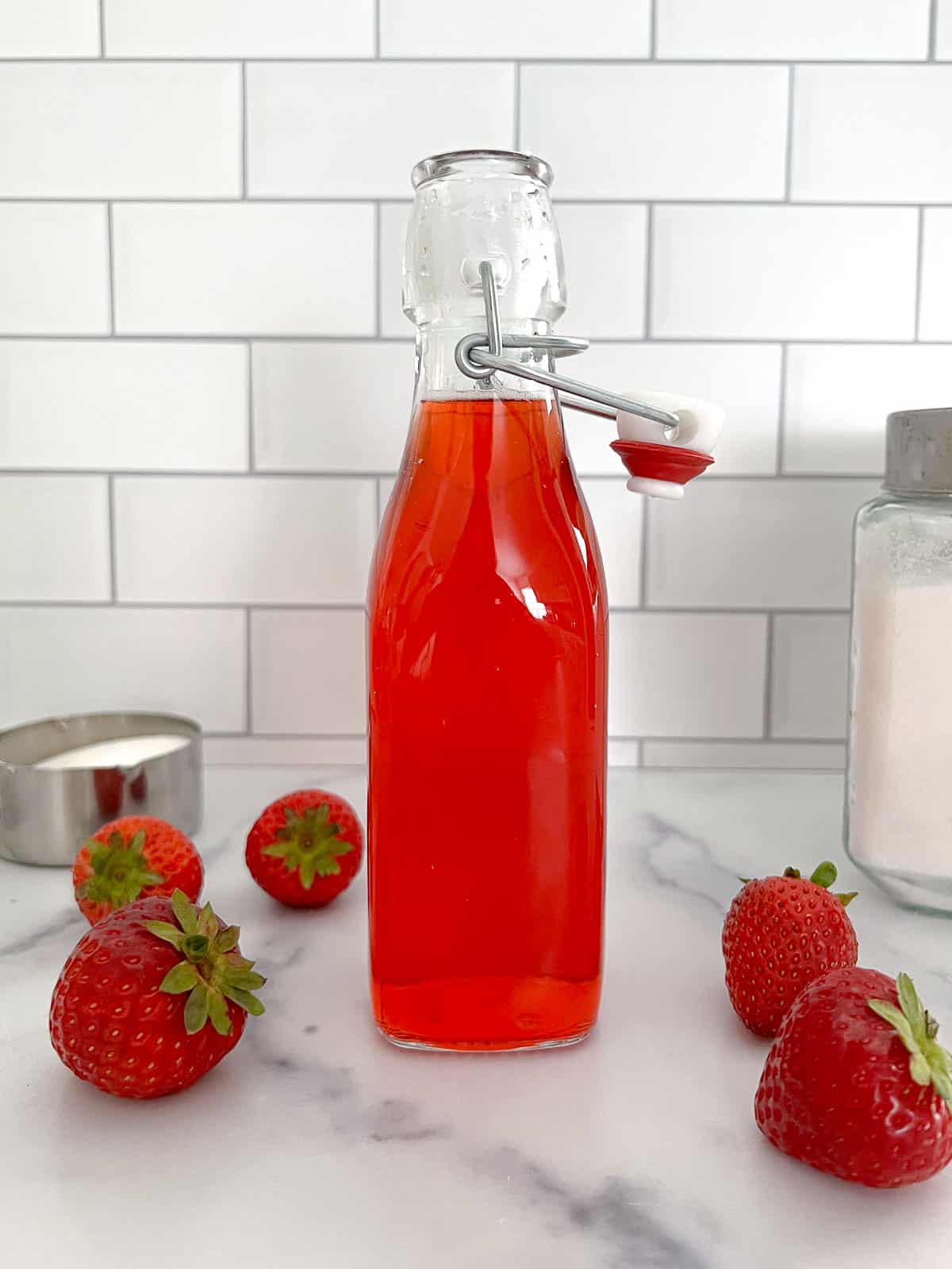 Homemade strawberry simple syrup in a glass bottle with open lid and sugar on the side.
