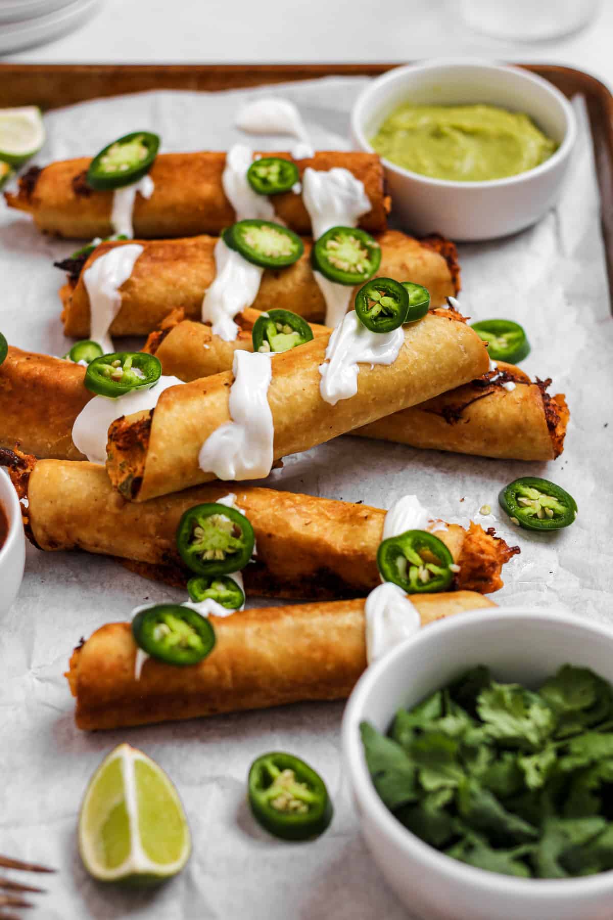 Chicken flautas on baking sheet with sour cream and jalapenos.