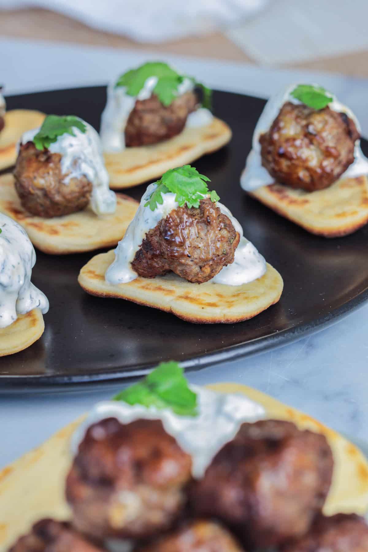 Lamb meatballs over pita bread on black plate with sauce.