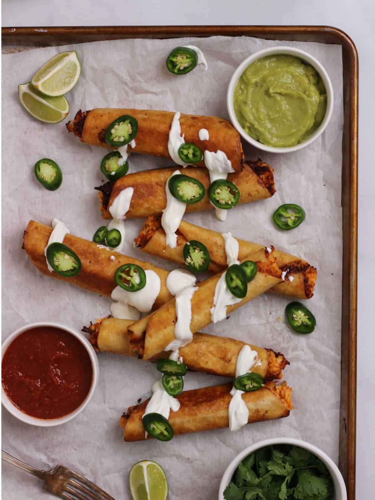 Chicken flautas drizzled with sour cream and jalapenos on a sheet pan.