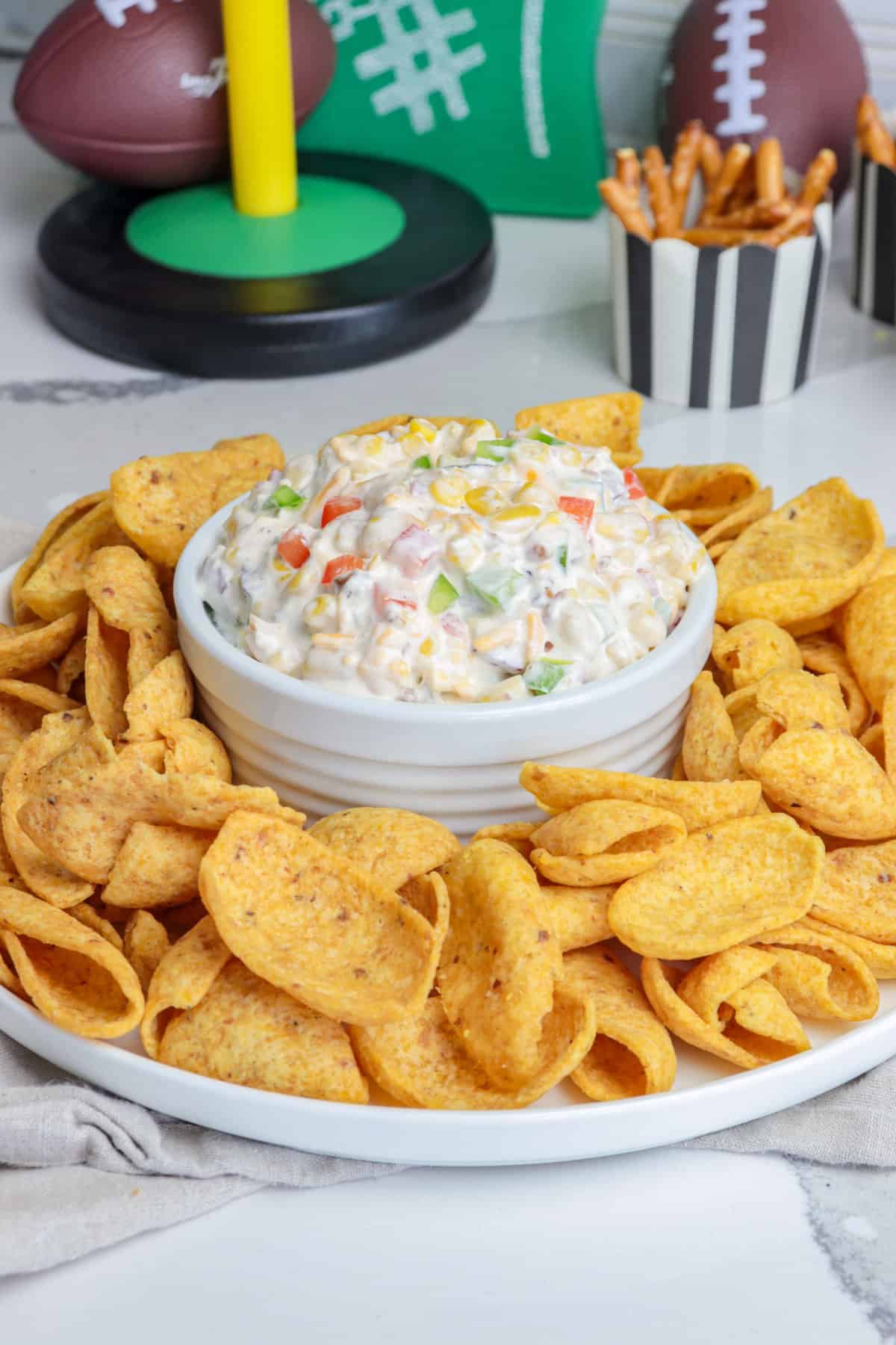 A white bowl with a corn crack dip with peppers, corn and cheeses surrounded by Fritos.