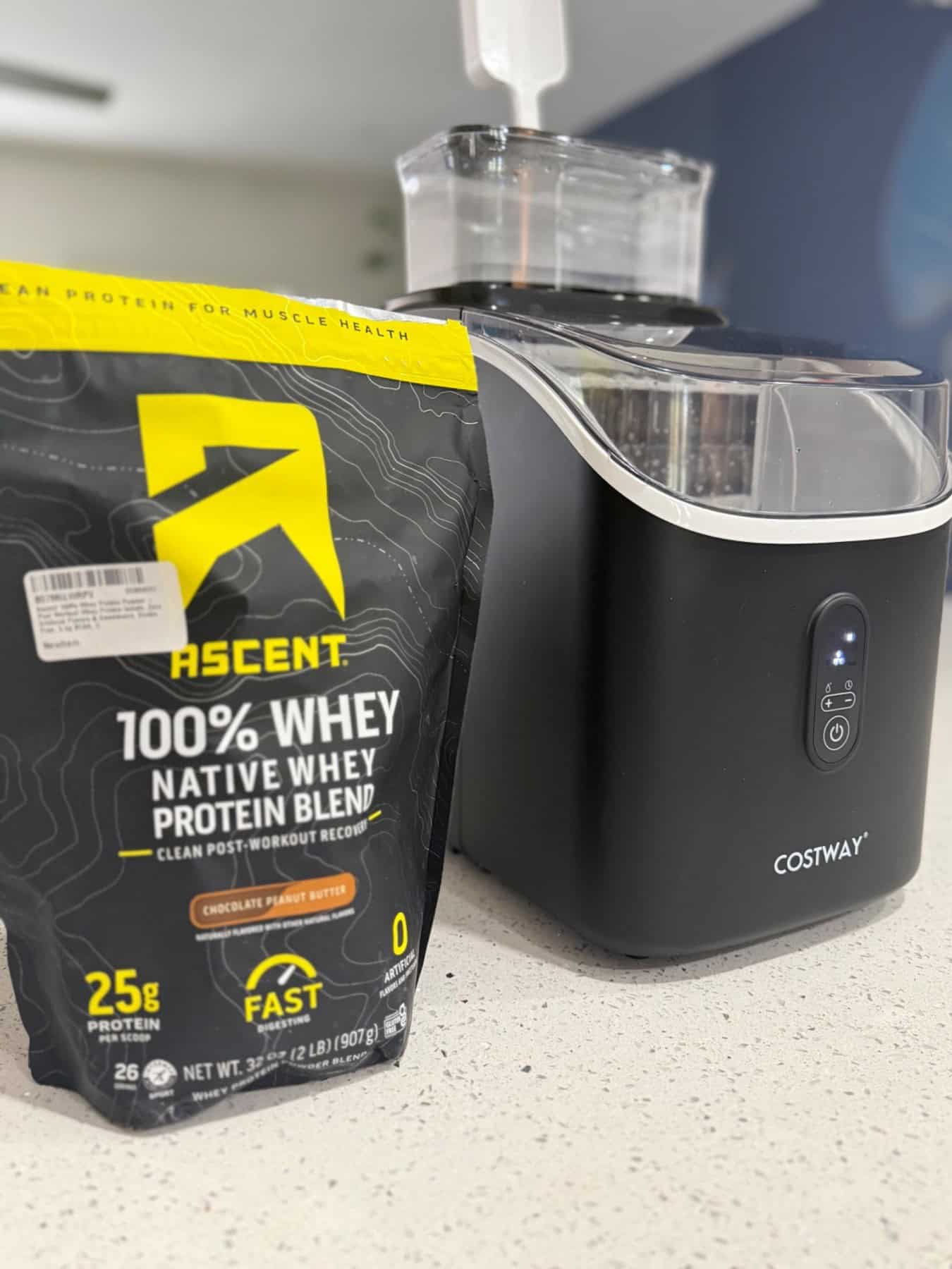 Black COSTWAY countertop ice maker beside a protein bag for size comparison on a kitchen counter.